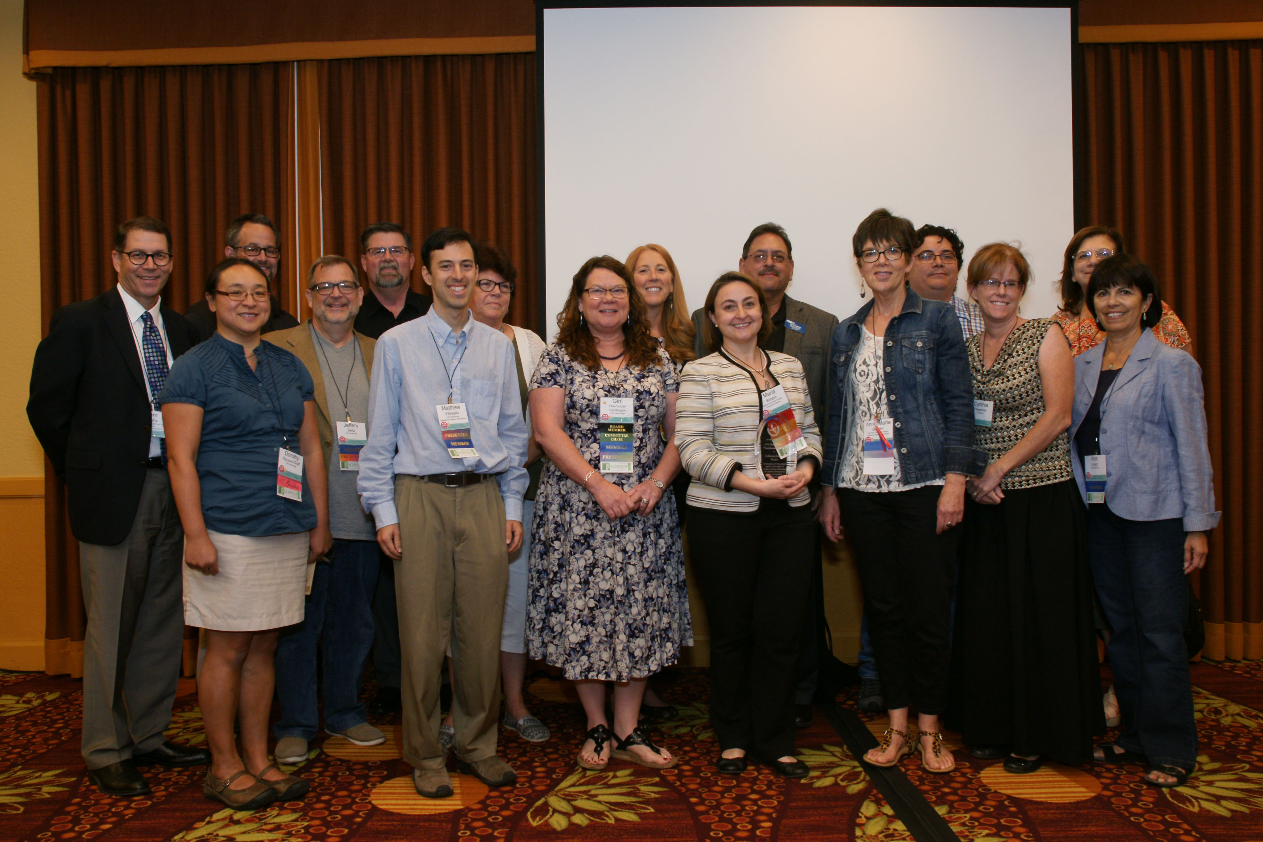 Sixteen California Science educators in semi-formal attire wearing conference lanyards are posing and  standing together and smiling in a conference room.