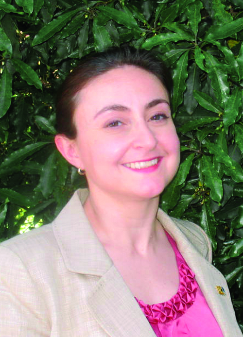A woman with brown hair, a pink shirt and tan blazer, Maria Simani, is standing in front of green leaves.